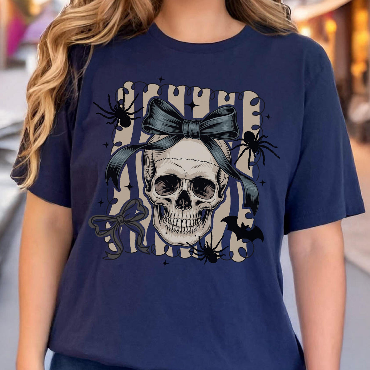 May include: Navy blue t-shirt featuring a skull design with a dark blue bow, spiders, and a bat. The skull is centered on a striped background. A bow is also on the left side of the skull. The shirt is a casual wear item.