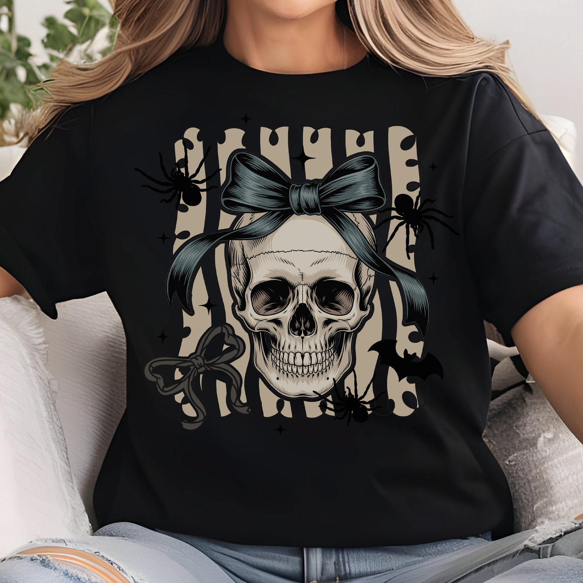 May include: Black t-shirt featuring a skull with a dark blue bow, spiders, and bats. The skull is centered on a striped background. A second bow is below the skull. The design has a Halloween theme.