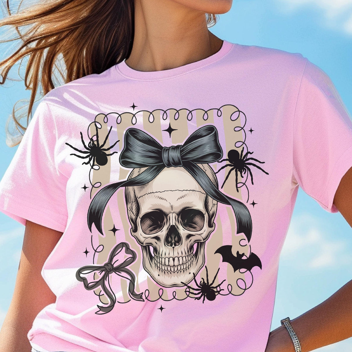 May include: A light pink t-shirt featuring a skull with a black bow, spiders, and a bat. The skull has a black bow tied around it. The design is surrounded by a decorative frame with stars and swirls. A Halloween-themed graphic tee.