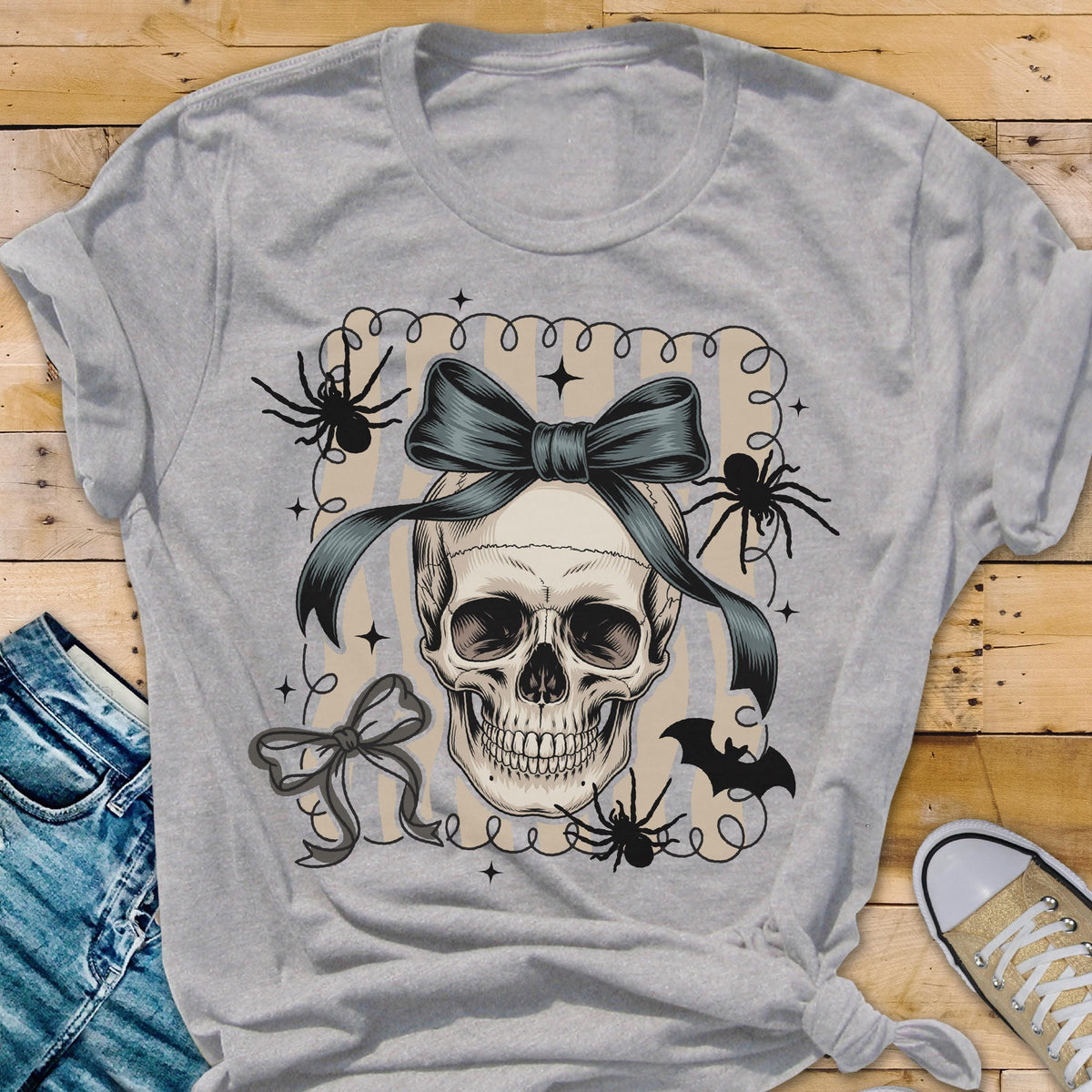 May include: A light gray t-shirt featuring a skull with a teal bow, spiders, and a bat. The skull is the central design element, adorned with a large bow. Spiders and a bat are placed around the skull, creating a Halloween theme.