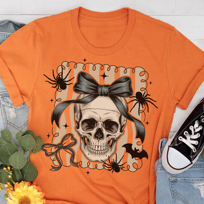 May include: Orange t-shirt featuring a skull with a black bow, spiders, and a bat. The skull is centered on a striped background with decorative swirls and stars. A black and white sneaker is visible on the right side.