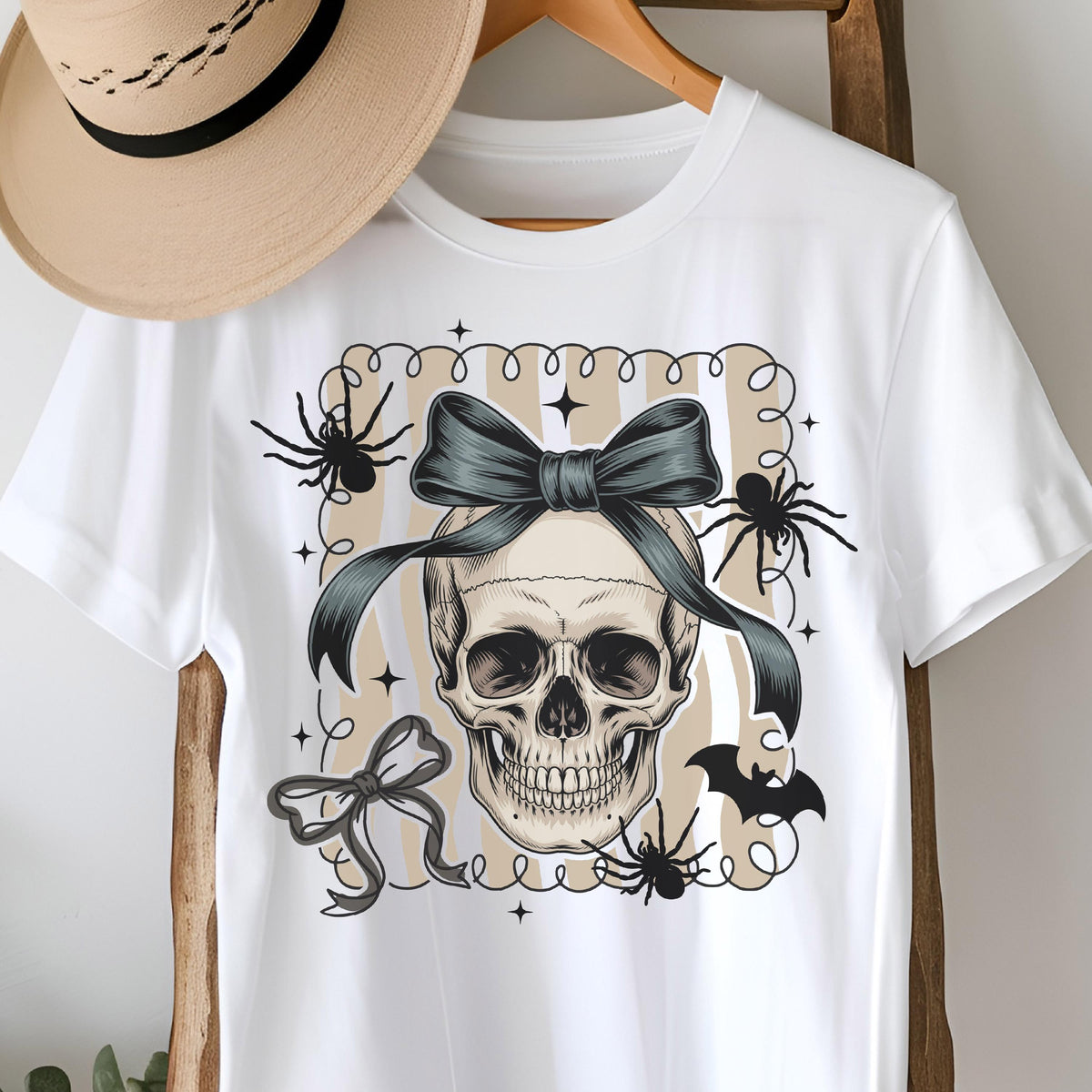 May include: White t-shirt featuring a skull with a teal bow, spiders, and a bat. The design is set within a beige and white striped frame. The shirt is displayed on a wooden hanger.