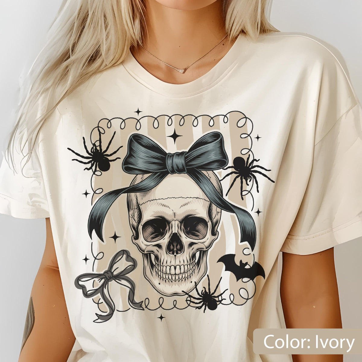 May include: Ivory-colored t-shirt featuring a detailed skull graphic with a dark blue bow, spiders, and a bat. The design is framed with a striped background and decorative elements, perfect for Halloween or gothic themes.