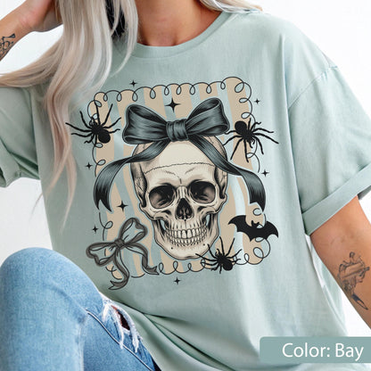 May include: A light blue t-shirt featuring a skull with a dark bow, spiders, and a bat graphic. The design includes a decorative frame with stars and a smaller bow. The shirt's color is bay.
