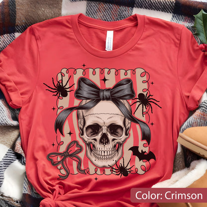 May include: Crimson-colored t-shirt featuring a skull with a black bow, spiders, and a bat graphic. The design is set within a striped frame. The shirt is made of a soft material.