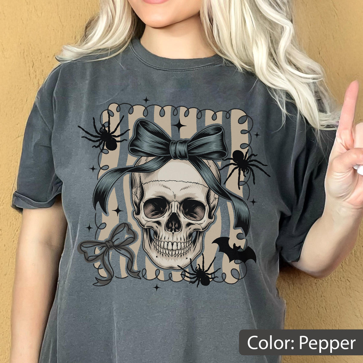 May include: Pepper-colored t-shirt featuring a skull with a dark blue bow, spiders, and a bat graphic. The design is set against a striped background with a decorative border. The shirt is a relaxed fit.