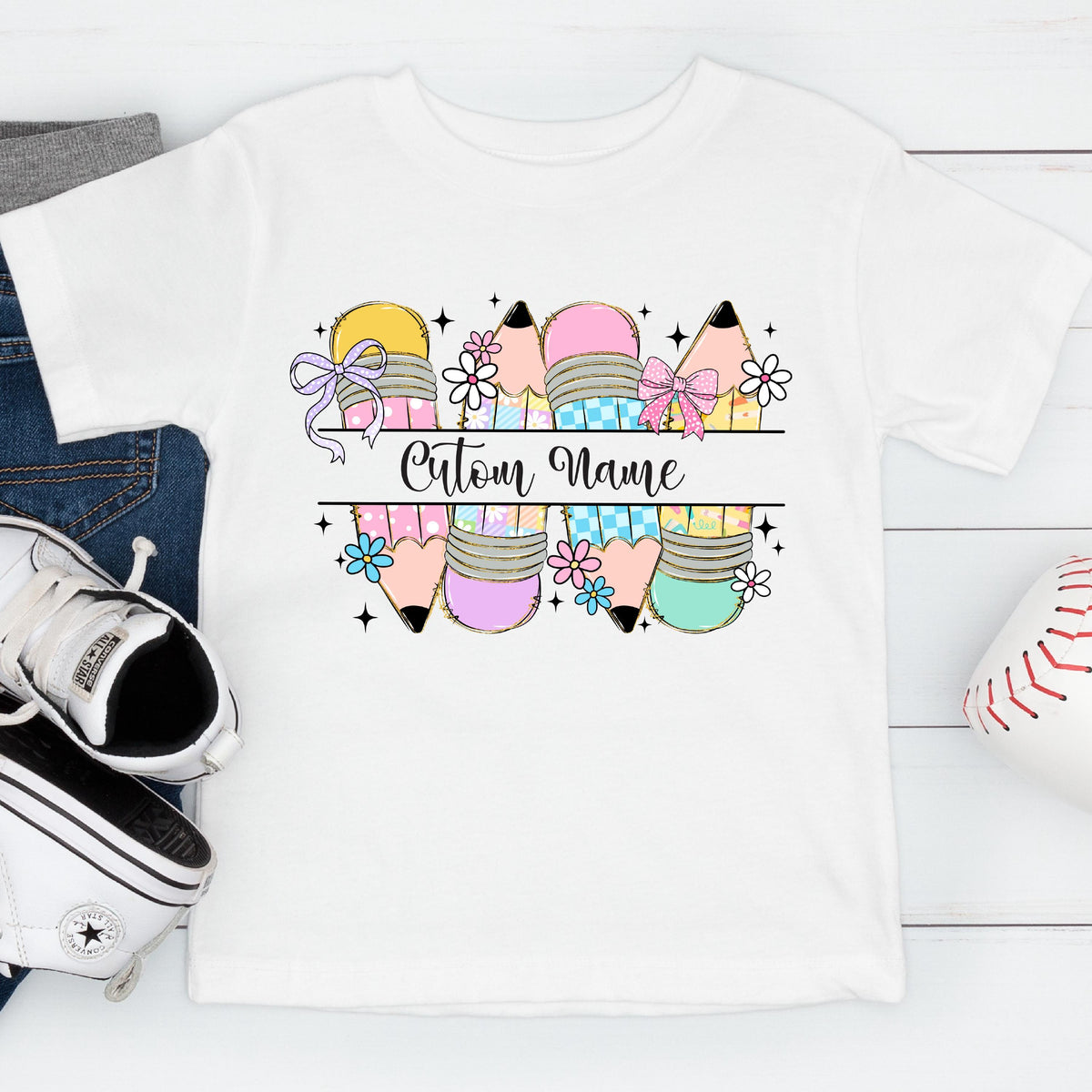 May include: White t-shirt featuring a colorful pencil design with bows and flowers. The design includes the text "Cutour Name" in a decorative font, perfect for personalization. The shirt is paired with blue jeans, a white sneaker, and a baseball.