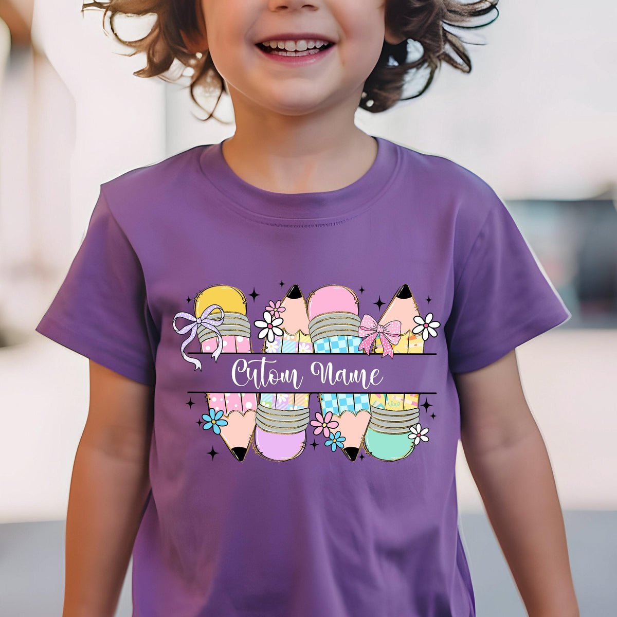 May include: A purple t-shirt featuring a colorful pencil design with the text "Custom Name". The pencils have various colors, bows, and floral accents. The shirt is worn by a child.