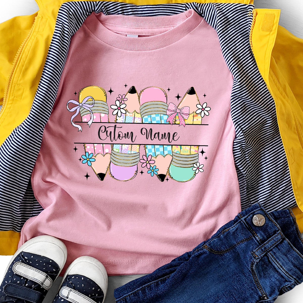 May include: Pink t-shirt with a pencil design and the text "Custom Name". The pencils are in yellow, pink, blue, and green with bows and flowers. The shirt is paired with blue jeans, a yellow jacket, and sneakers.