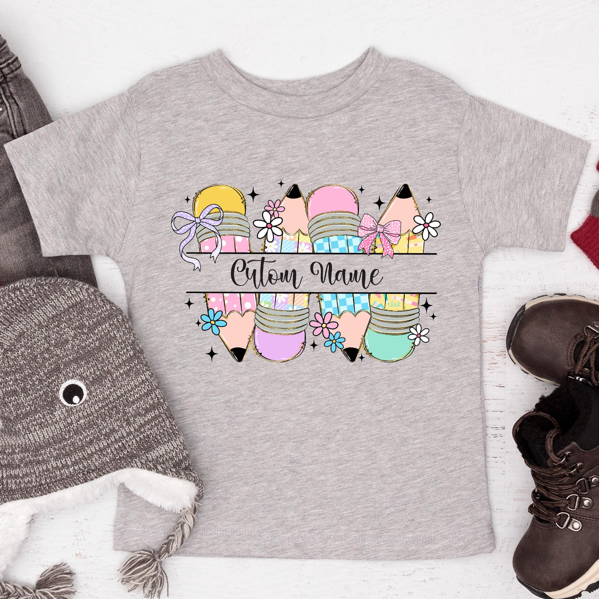 May include: A light gray toddler t-shirt featuring a colorful pencil design with the text "Custom Name". The pencils are in yellow, pink, blue, and green, with floral and bow accents. A cute and playful design for kids.