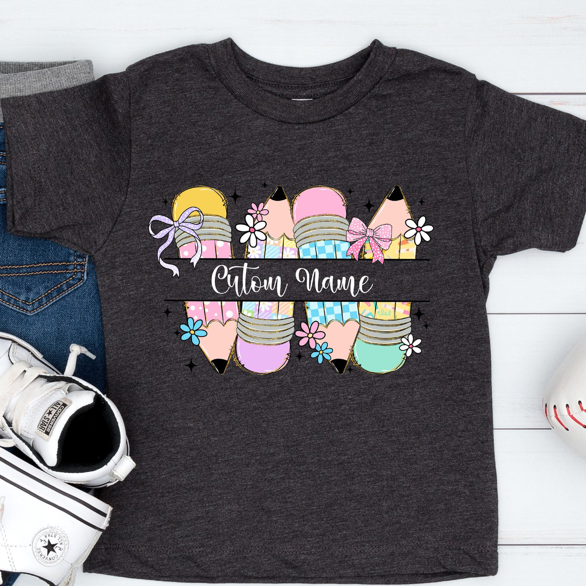 May include: Dark gray t-shirt featuring a colorful pencil design with the text "Custom Name". The pencils are in shades of yellow, pink, purple, and teal, decorated with bows and flowers. A fun and personalized tee for kids.
