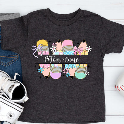 May include: Dark gray t-shirt featuring a colorful pencil design with the text "Custom Name". The pencils are in shades of yellow, pink, purple, and teal, decorated with bows and flowers. A fun and personalized tee for kids.