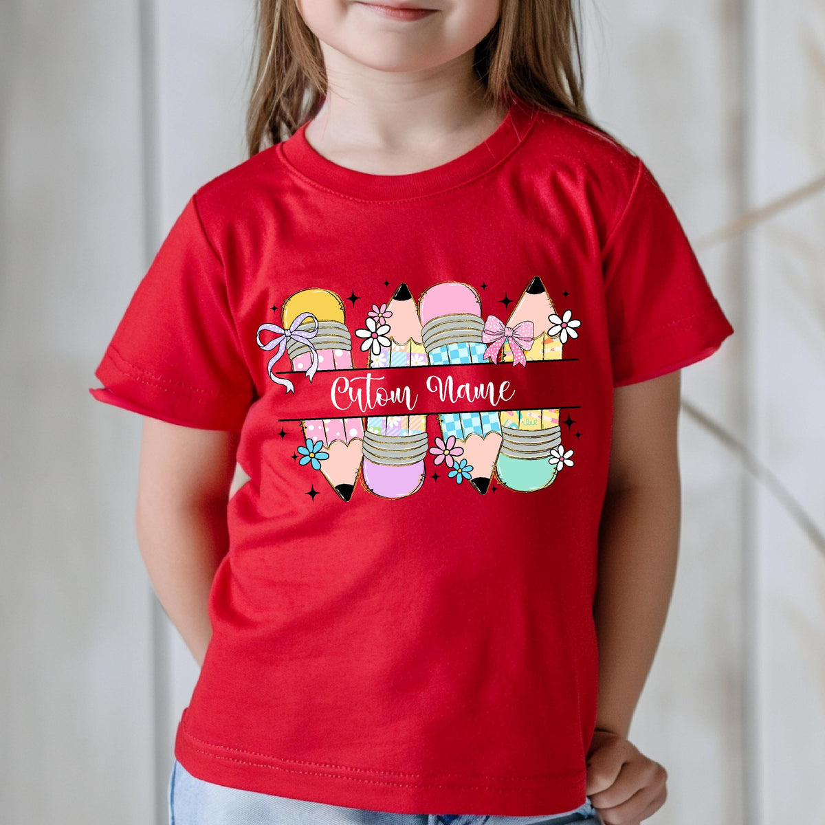 May include: A red t-shirt featuring a colorful pencil design with the text "Custom Name" in a cursive font. The pencils have various colors, bows, and floral accents, creating a playful and personalized look.