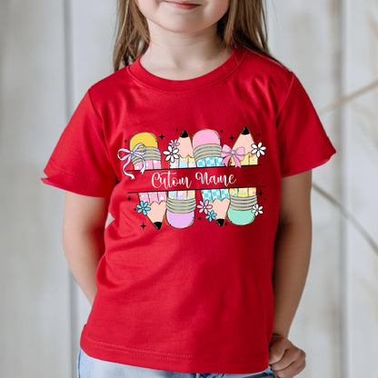May include: A red t-shirt featuring a colorful pencil design with the text "Custom Name" in a cursive font. The pencils have various colors, bows, and floral accents, creating a playful and personalized look.