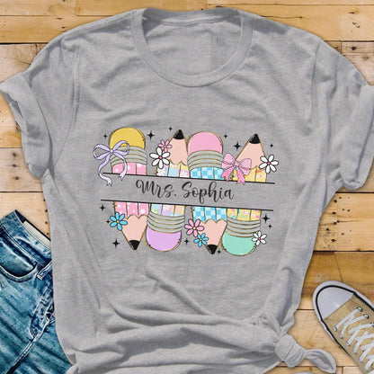 May include: A gray t-shirt featuring a colorful pencil design with bows and flowers. The text "Mrs. Sophia" is displayed in a cursive font. The pencils are in various pastel colors, perfect for a teacher.