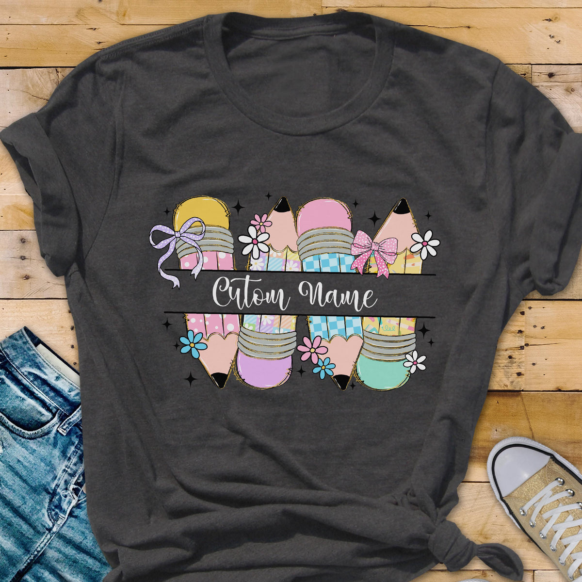 May include: Dark gray t-shirt featuring a colorful pencil design with the text "Custom Name". The pencils are decorated with bows and flowers in pastel colors. A personalized gift for teachers or students.