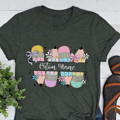 May include: Dark gray t-shirt featuring a colorful pencil design with the text "Custom Name". The pencils are in various colors, including yellow, pink, and blue, with floral and bow accents. A cute and fun design for teachers or students.