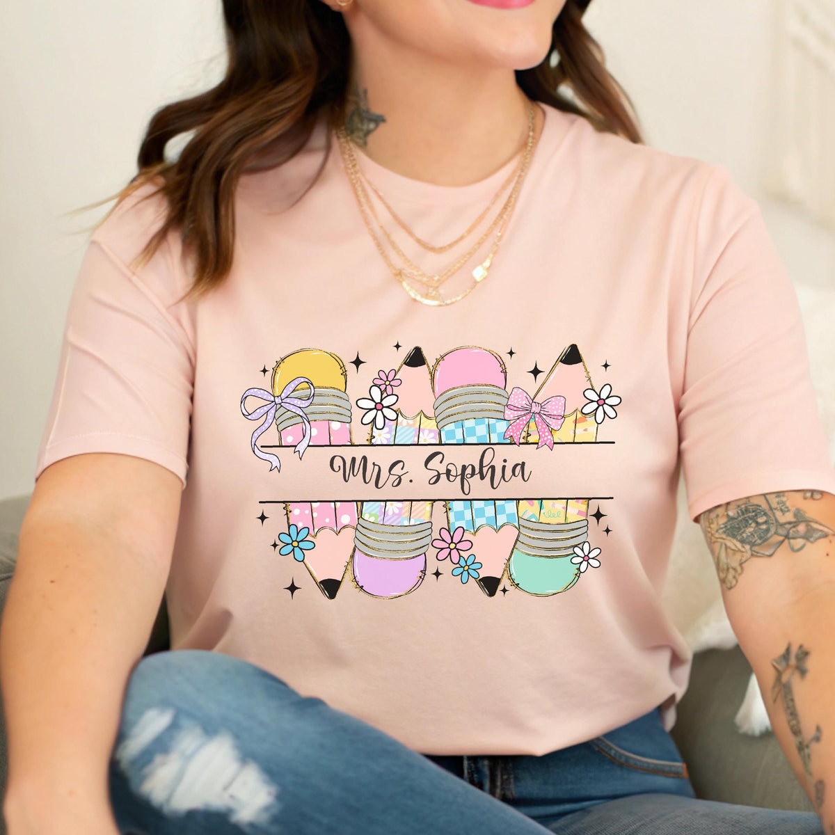 May include: A light pink t-shirt featuring a colorful pencil design with the text "Mrs. Sophia". The pencils are decorated with bows and flowers. The shirt is a crew neck style.