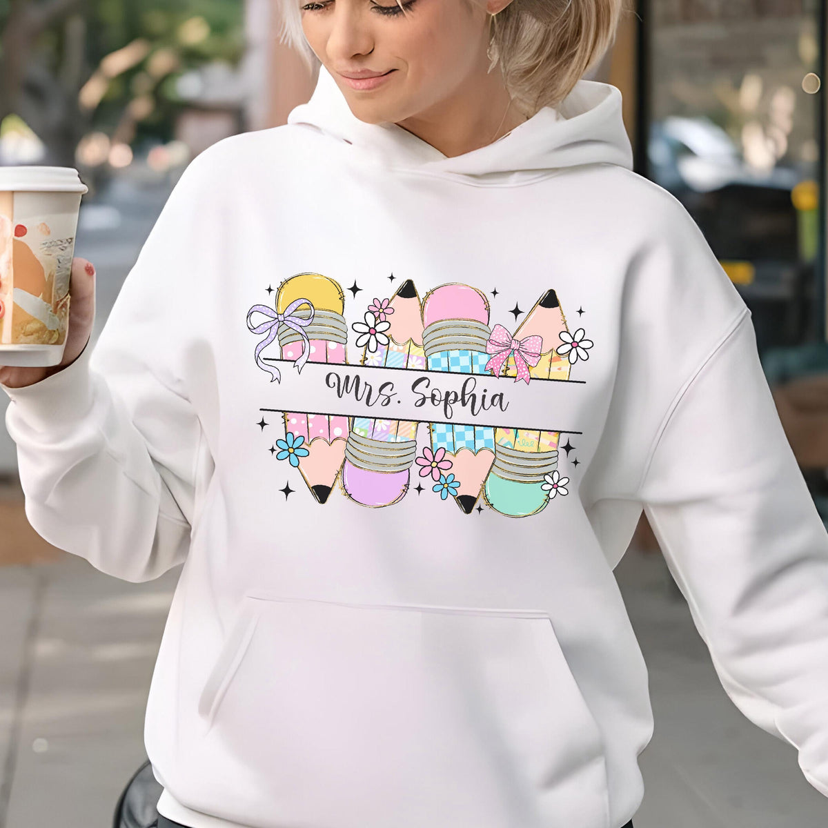 May include: White hoodie with a colorful pencil design. The design includes pencils in yellow, pink, and teal, with bows and flowers. The text "Mrs. Sophia" is printed in a script font.
