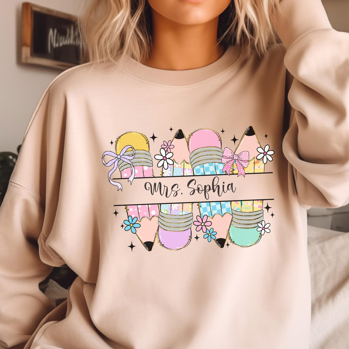 May include: Beige sweatshirt featuring a colorful pencil design with the text "Mrs. Sophia". The pencils are decorated with bows and flowers. The design includes yellow, pink, blue, and green colors.