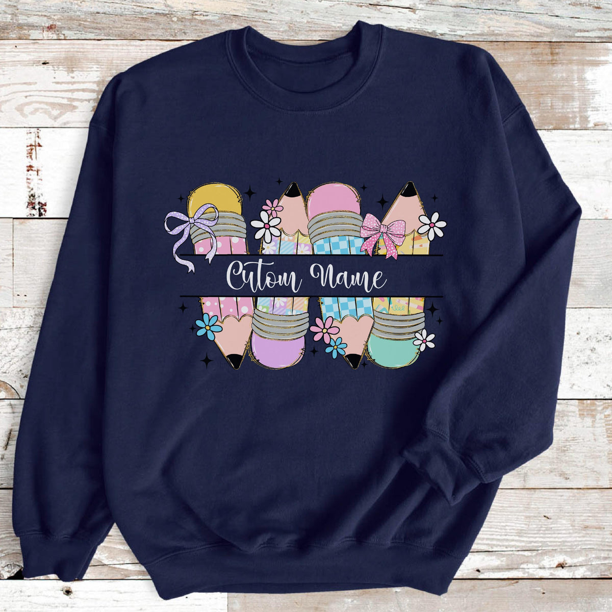 May include: Navy blue sweatshirt featuring a pencil design with pastel colors and floral accents. The pencils are decorated with bows and the text "Custom Name" is displayed across the center. A cute and colorful design.