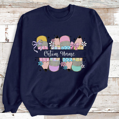 May include: Navy blue sweatshirt featuring a pencil design with pastel colors and floral accents. The pencils are decorated with bows and the text "Custom Name" is displayed across the center. A cute and colorful design.