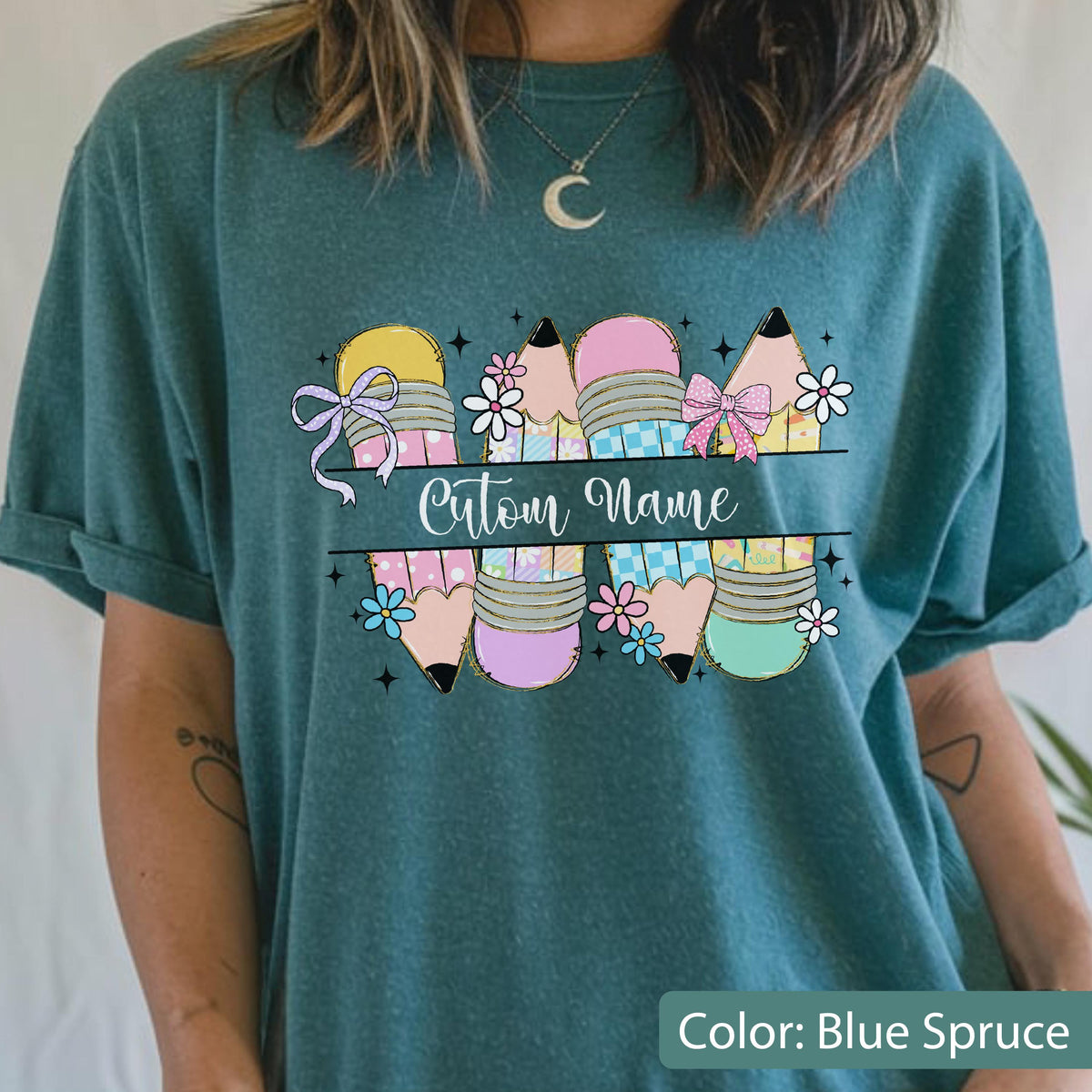 May include: A blue spruce t-shirt featuring a graphic of four pencils in pastel shades of pink, yellow, and blue, decorated with small flowers and bows. A horizontal line across the center contains the text Cutom Name in white cursive script.