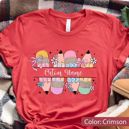 May include: A crimson short-sleeved t-shirt featuring a graphic of four pencils with pastel patterns, pink bows, and small flowers. A horizontal line divides the pencils, with the text "Cutom Name" written in white cursive script in the center.