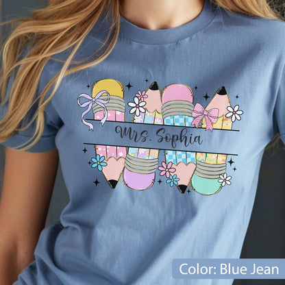 May include: A blue jean colored t-shirt featuring a graphic of four pencils with pastel patterns, bows, and small flowers. A horizontal black line divides the pencils, with the text Mrs. Sophia written in a cursive black script across the center.