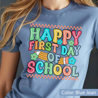 May include: A blue jean colored t-shirt featuring a graphic print with the text HAPPY FIRST DAY OF SCHOOL in colorful, dotted bubble letters. The design includes a yellow pencil illustration, small flowers, and orange checkered borders.