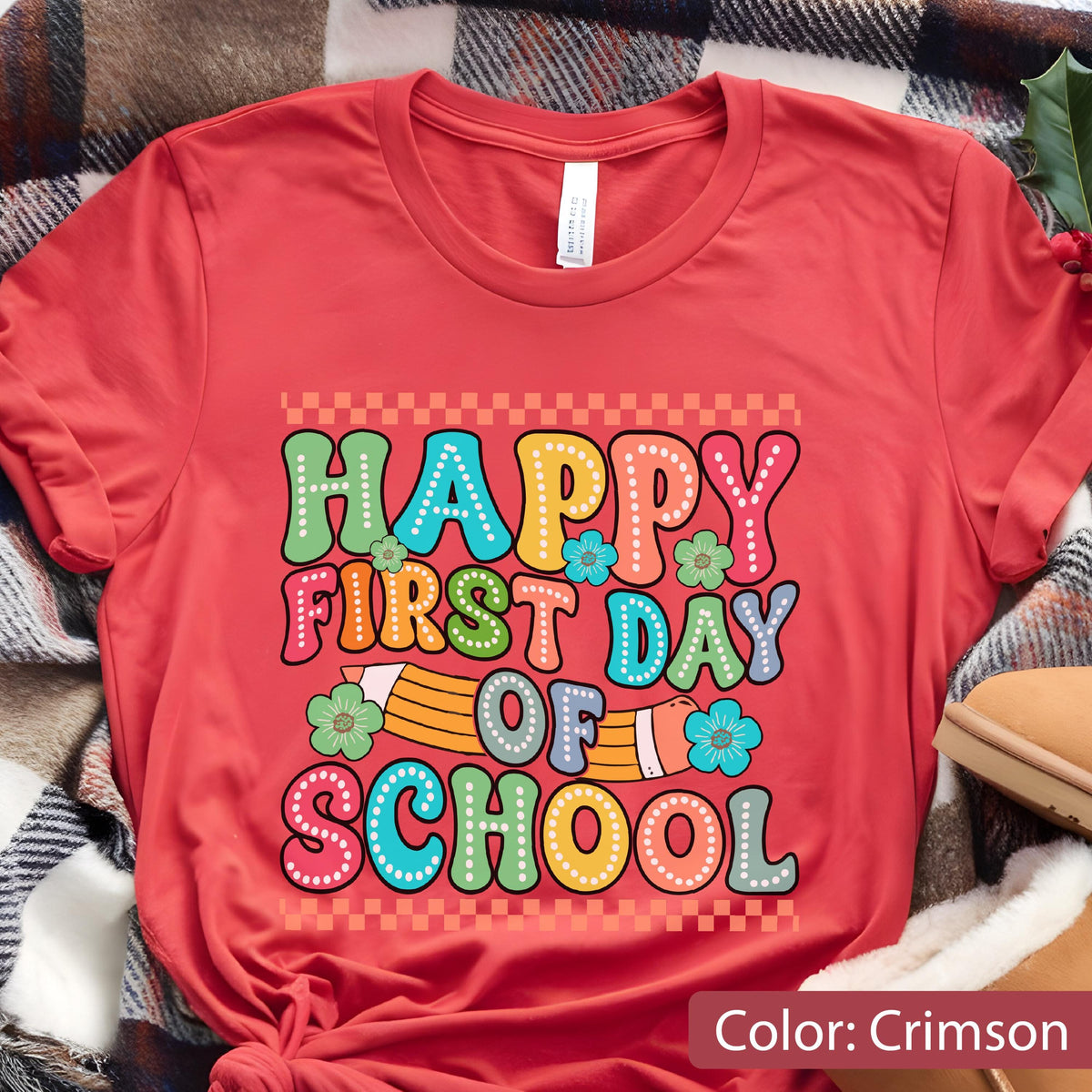 May include: A crimson short-sleeved t-shirt featuring a graphic print with the text "HAPPY FIRST DAY OF SCHOOL" in colorful, bubbly letters. The design includes a yellow pencil illustration, small green and blue flowers, and a checkered border.