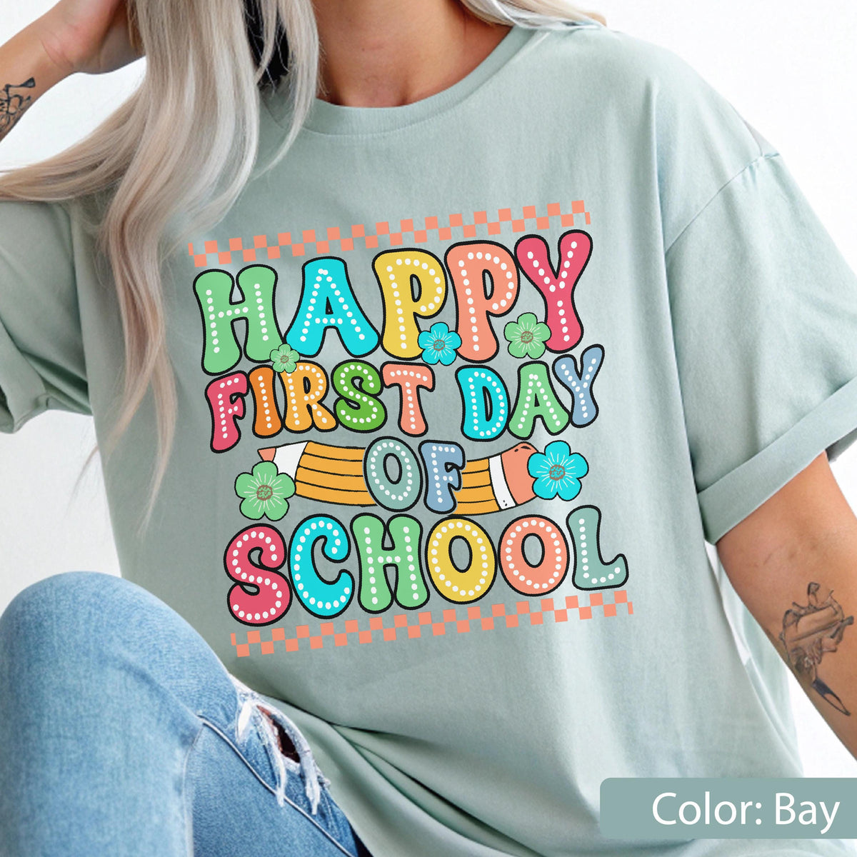 May include: A bay-colored t-shirt featuring a graphic print with the text "HAPPY FIRST DAY OF SCHOOL" in colorful, dotted bubble letters. The design includes a yellow pencil illustration, small blue and green flowers, and orange checkered borders.