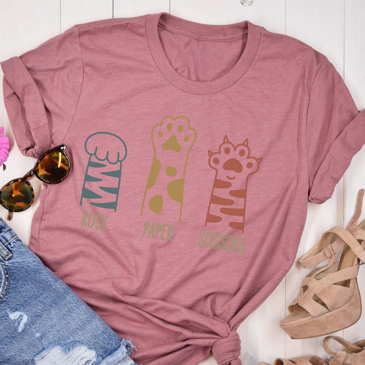 May include: A mauve short-sleeved t-shirt featuring a graphic of three animal paws with the text Rock, Paper, Scissors printed below each paw. The first paw is teal with stripes, the second is gold with spots, and the third is brown with stripes.