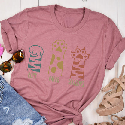 May include: A mauve short-sleeved t-shirt featuring a graphic of three animal paws with the text Rock, Paper, Scissors printed below each paw. The first paw is teal with stripes, the second is gold with spots, and the third is brown with stripes.