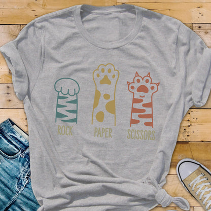 May include: A light gray short-sleeve t-shirt featuring a graphic of three cat paws representing the game rock, paper, scissors. The first paw is teal with stripes, the second is gold with spots, and the third is orange with stripes and text below.