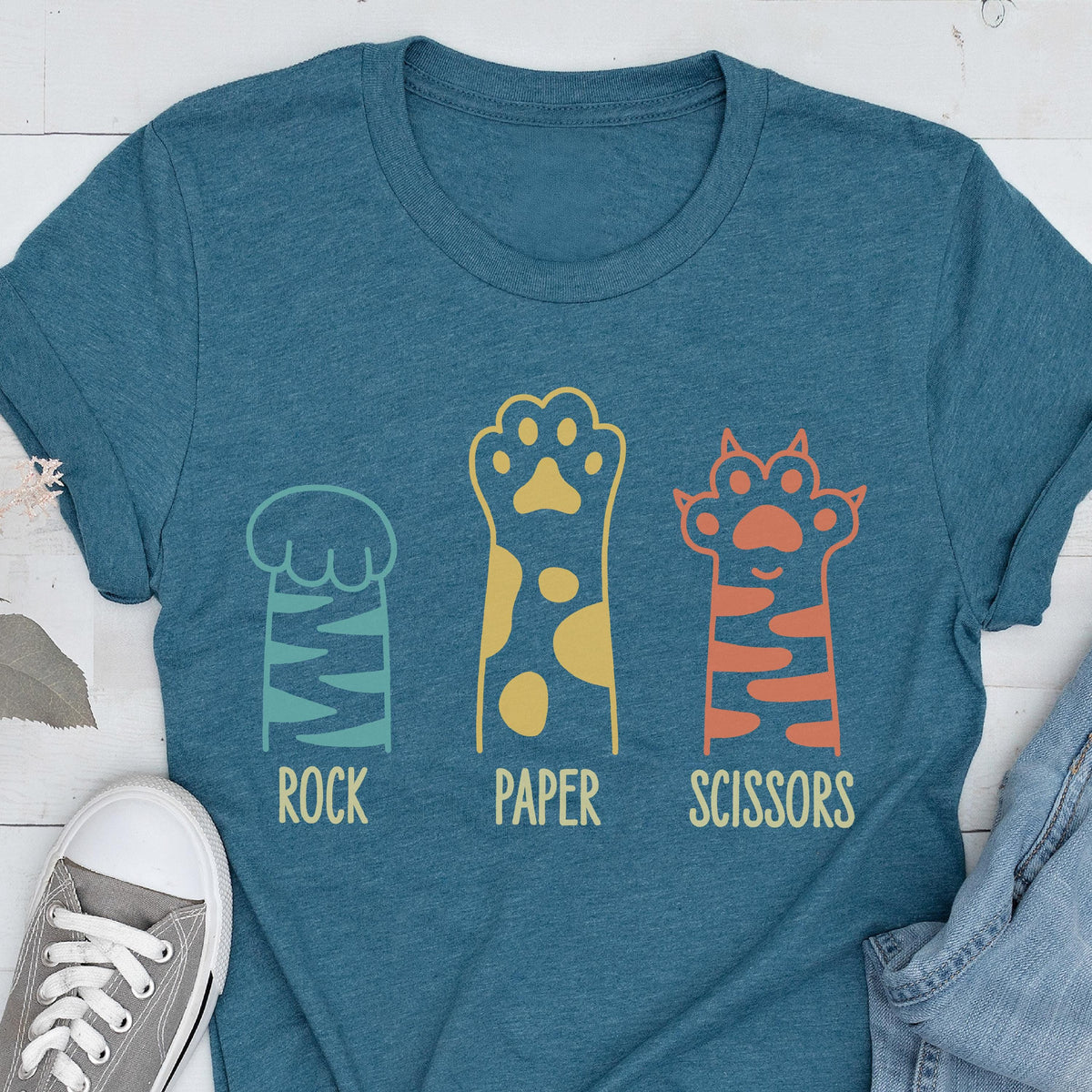 May include: A heathered teal short-sleeve t-shirt featuring a graphic of three animal paws. The first paw is light blue with stripes labeled ROCK, the second is yellow with spots labeled PAPER, and the third is orange with stripes labeled SCISSORS.