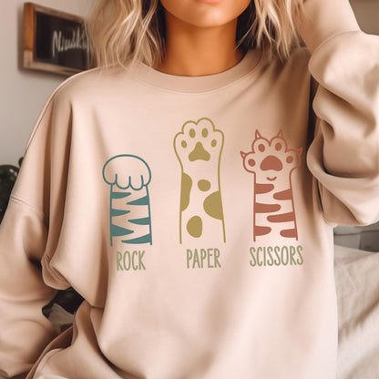 May include: A beige crewneck sweatshirt featuring a graphic of three animal paws representing the game Rock, Paper, Scissors. The first paw is teal with stripes, the second is yellow with spots, and the third is terracotta with stripes and claws.