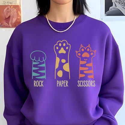 May include: A vibrant purple crewneck sweatshirt featuring a graphic print of three animal paws arranged in a row. Below each paw, the text reads ROCK, PAPER, and SCISSORS in a light yellow font. The paws are colored in teal, yellow, and orange.