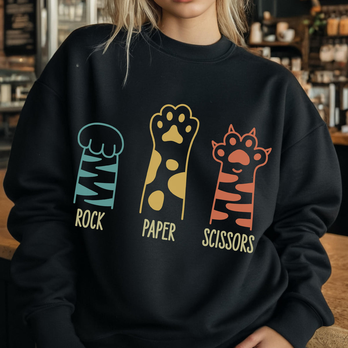 May include: A black long-sleeve sweatshirt featuring a graphic of three animal paws. The first paw is teal with stripes, labeled ROCK. The second is yellow with spots, labeled PAPER. The third is orange with stripes, labeled SCISSORS.
