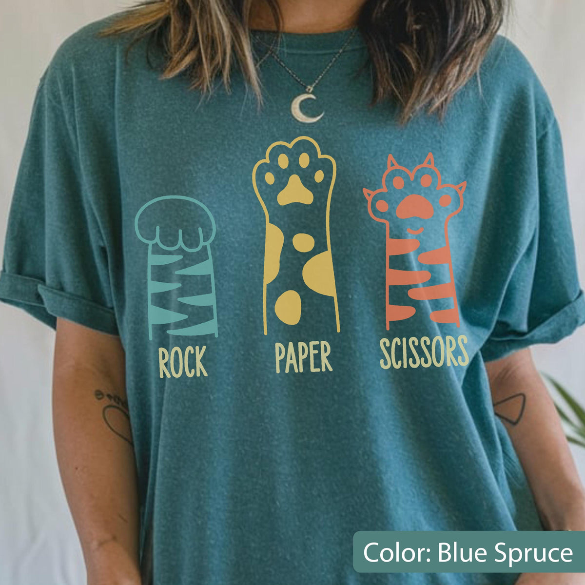 May include: A blue spruce t-shirt featuring a graphic of three animal paws representing rock, paper, and scissors. The first paw is teal with stripes, the second is yellow with spots, and the third is orange with stripes, with text below each.