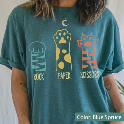May include: A blue spruce t-shirt featuring a graphic of three animal paws representing rock, paper, and scissors. The first paw is teal with stripes, the second is yellow with spots, and the third is orange with stripes, with text below each.
