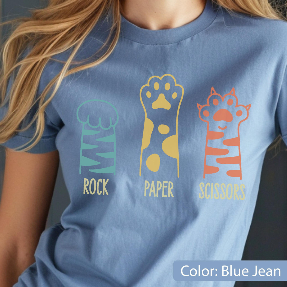 May include: A blue jean colored t-shirt featuring a graphic of three animal paws representing rock, paper, and scissors. The first paw is teal with stripes, the second is yellow with spots, and the third is orange with stripes. Text reads ROCK PAPER SCISSORS.