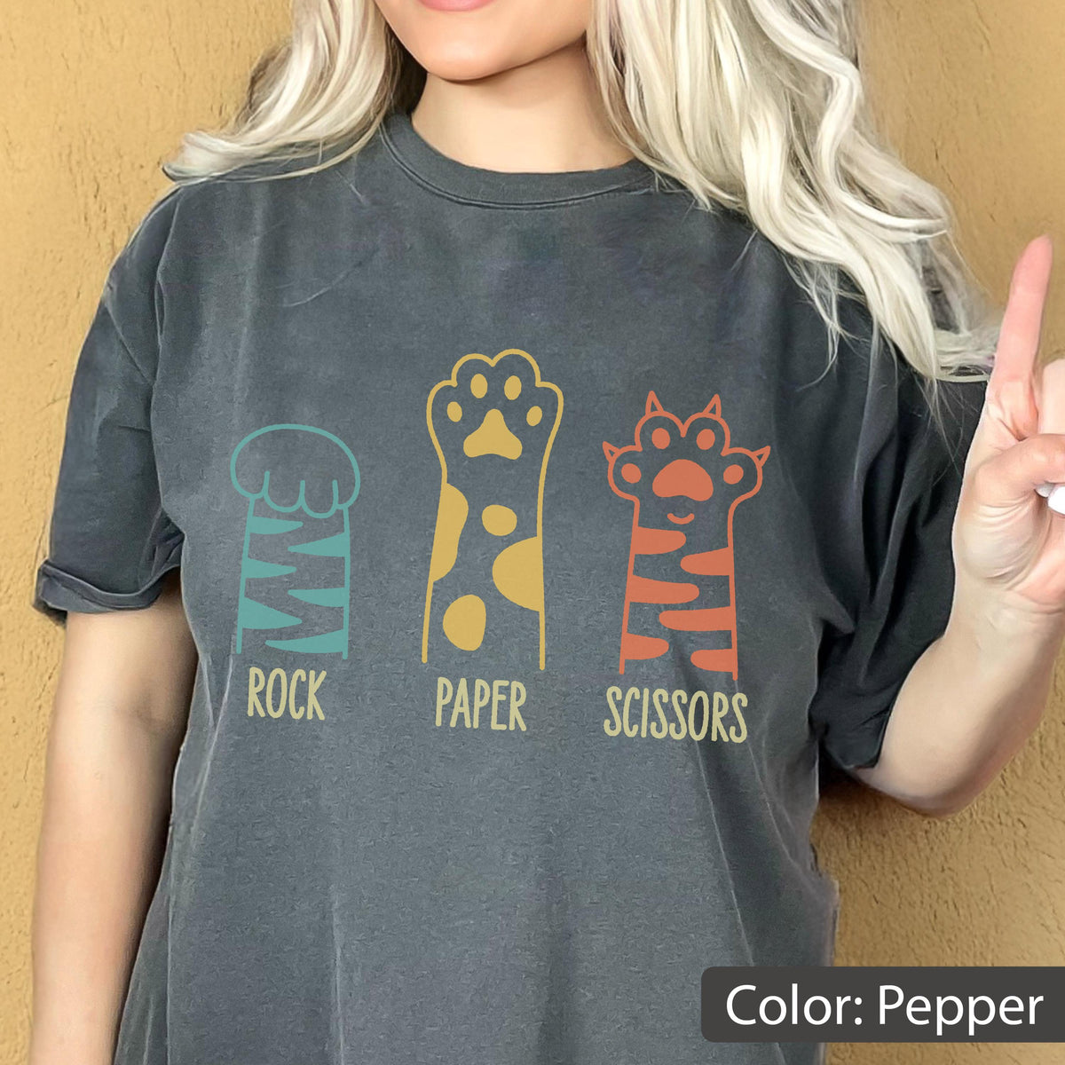 May include: A pepper-colored short-sleeve t-shirt featuring a graphic of three animal paws representing rock, paper, and scissors. The first paw is teal with stripes, the second is yellow with spots, and the third is orange with stripes.