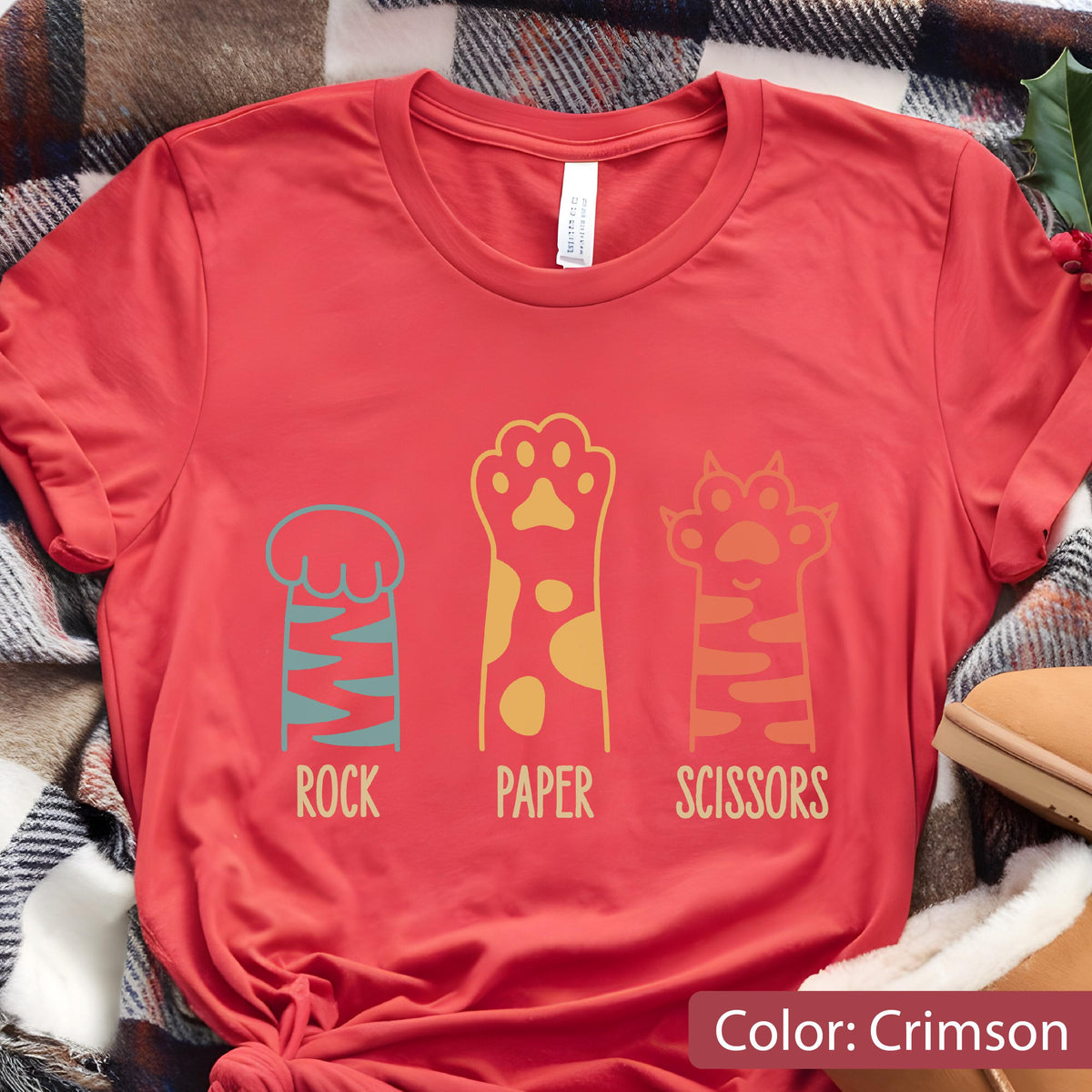 May include: A crimson short-sleeved t-shirt featuring a graphic of three animal paws representing the game rock, paper, scissors. The first paw is teal with stripes, the second is yellow with spots, and the third is orange with stripes.