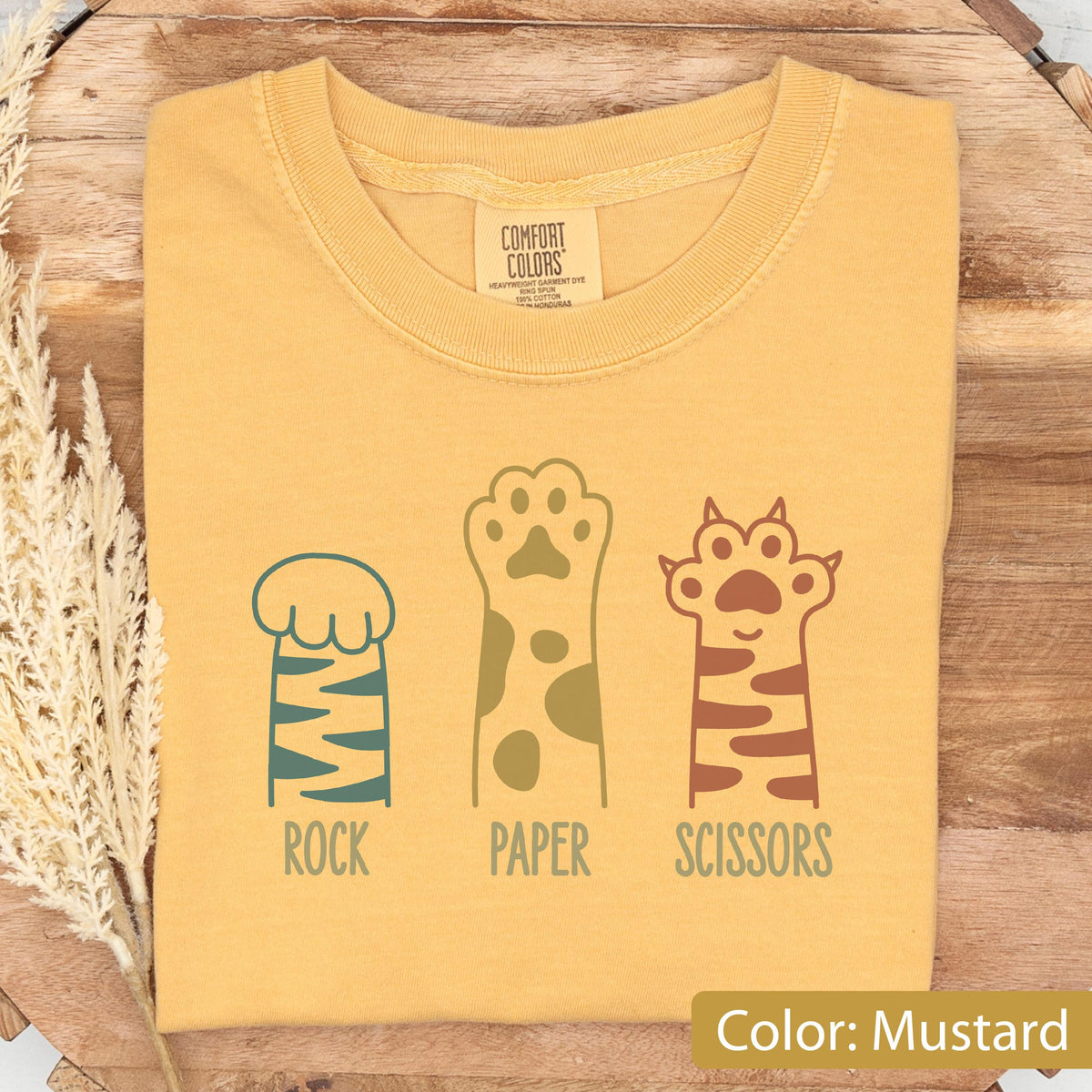 May include: A mustard yellow t-shirt featuring a graphic of three animal paws representing rock, paper, and scissors. The first paw is blue with stripes, the second is yellow with spots, and the third is brown with stripes. The text reads ROCK, PAPER, SCISSORS.