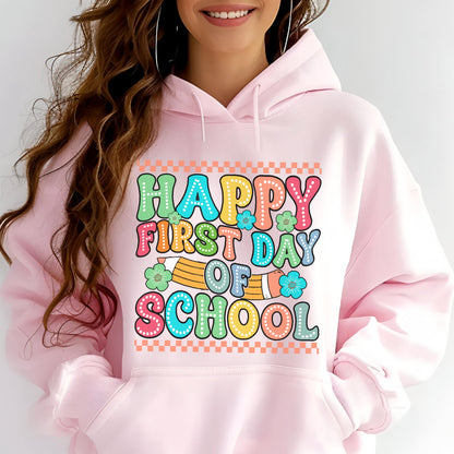 May include: A light pink hooded sweatshirt featuring a graphic print with the text "HAPPY FIRST DAY OF SCHOOL" in colorful, dotted bubble letters. The design includes a yellow pencil illustration, small green and blue flowers, and a checkered border.