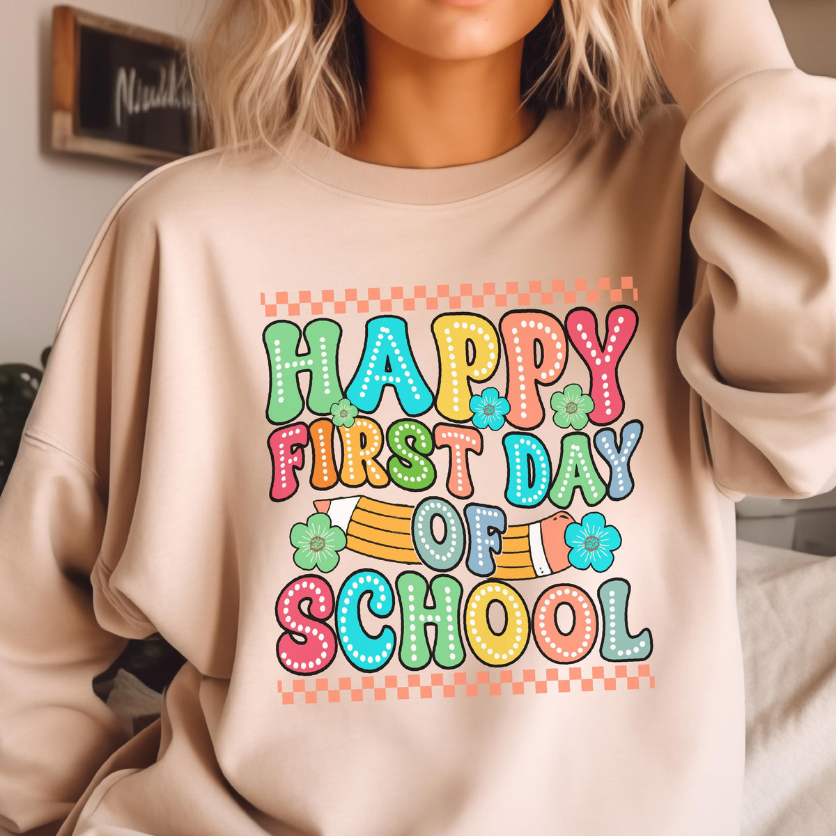 May include: A beige crewneck sweatshirt featuring a colorful graphic print that reads "HAPPY FIRST DAY OF SCHOOL" in bubble letters. The design includes a yellow pencil, small green and blue flowers, and a checkered border at the top and bottom.