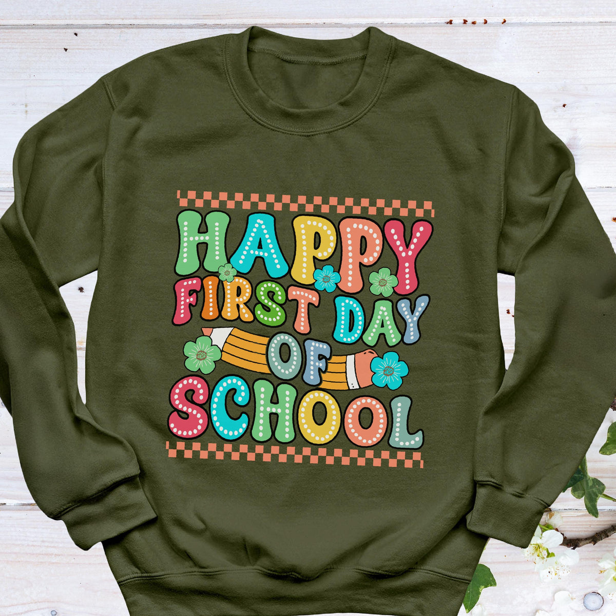 May include: An olive green crewneck sweatshirt featuring a colorful graphic print that reads Happy First Day Of School in bubble letters. The design includes a yellow pencil illustration, small flower accents, and orange checkered borders.