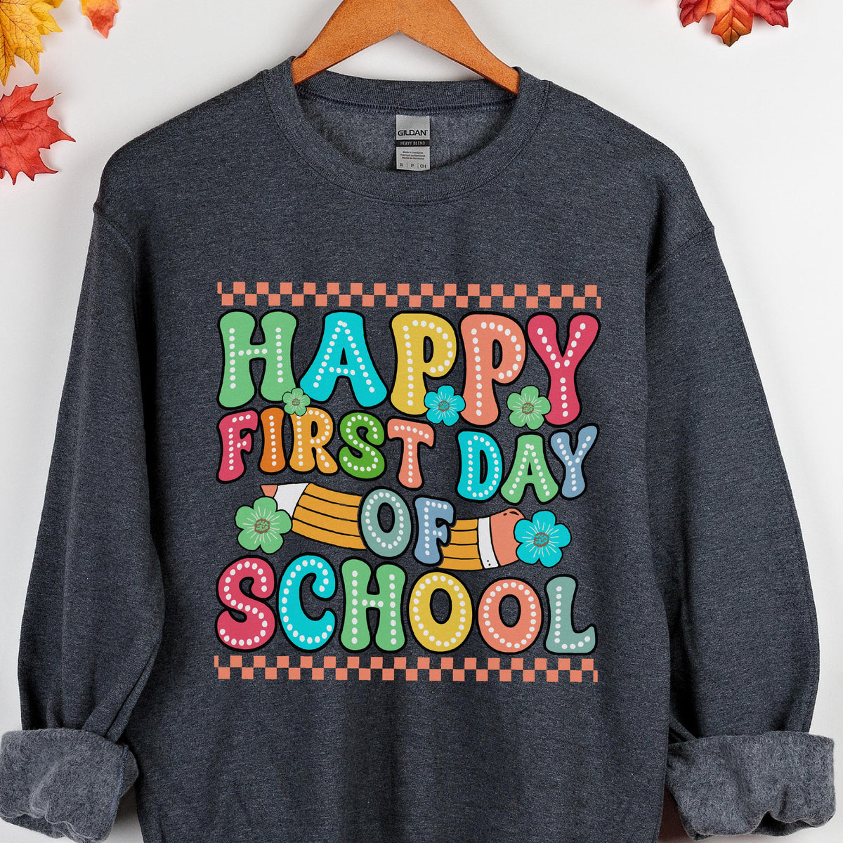 May include: A dark charcoal gray crewneck sweatshirt featuring a colorful graphic print that reads "HAPPY FIRST DAY OF SCHOOL" in a retro bubble font. The design includes a yellow pencil illustration, small flower accents, and a checkered border.