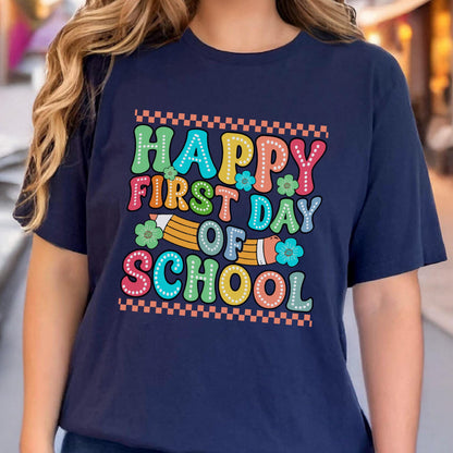 May include: A navy blue short-sleeved t-shirt featuring the text "HAPPY FIRST DAY OF SCHOOL" in a multi-colored, dotted, retro-style font. The design includes a yellow pencil graphic, small flower illustrations, and a checkered border pattern.